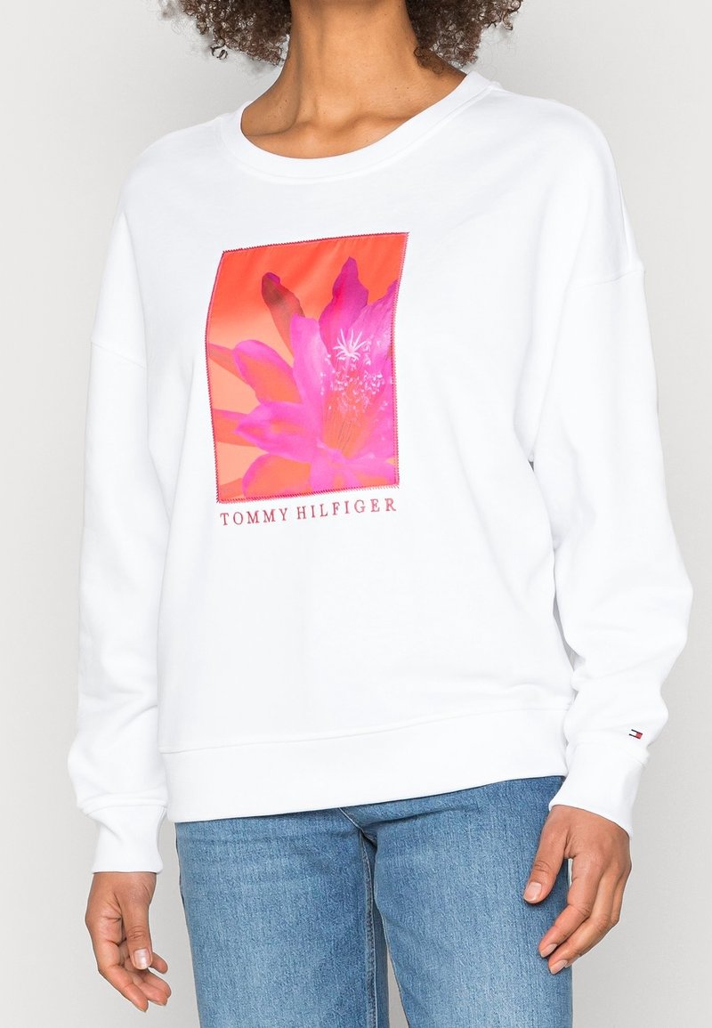 Sweatshirt blanc avec un graphique floral coloré sur le devant, le texte "TOMMY HILFIGER" en dessous, et des poignets et un ourlet côtelés. Tissu doux.