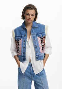 Denimweste mit gestickten Blumemustern in Rosa, Schwarz und Weiß. Verfügt über einen flauschigen Kragen und klassische Knopföffnung. Getragen über einem weißen Hemd.