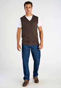 Brauner Strickwesten über einem weißen T-Shirt, kombiniert mit blauen Jeans und braunen Schnürschuhen. Die Weste hat ein V-Ausschnitt-Design.
