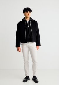 Νέος άνδρας με σγουρά μαλλιά, που φοράει μαύρο μπουφάν με φερμουάρ, λευκό παντελόνι και μαύρα παπούτσια, σηκωμένος μπροστά σε ένα απλό λευκό φόντο.