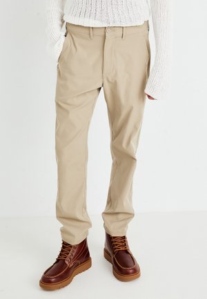 Personne portant un pantalon beige, un pull en laine blanc et des bottes en cuir marron à lacets, debout sur un sol blanc.