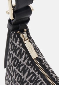 Torba DKNY z czarno-białym wzorem logo, teksturowaną tkaniną, złotym suwakiem oraz czarnym skórzanym paskiem z metalową klamrą.