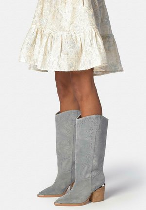 Botas grises de ante hasta la rodilla con patrones occidentales perforados y tacones gruesos de madera, combinadas con un vestido crema de encaje.