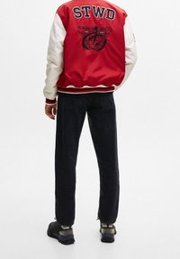 Veste varsity rouge et blanche avec un logo noir au dos, associée à un pantalon noir et des chaussures foncées. Comprend des poignets et un ourlet côtelés.