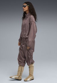 Bruine, glanzende stof jumpsuit met gathered details, hoge kraag en elastische manchetten, gecombineerd met beige kniehoge laarzen met veters en accenten.