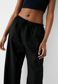 Kvinna i svart crop top och lösa svarta byxor med dragsko, sedd från midjan till övre låren mot enhällig bakgrund.