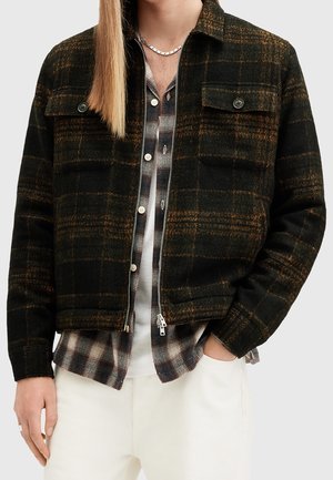 Veste en polaire à carreaux noirs et orange avec fermeture éclair sur le devant, deux poches poitrine et une coupe décontractée, portée par-dessus une chemise à carreaux.