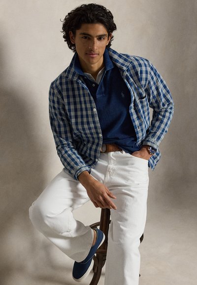 Young man seated on wooden stool wearing blue checkered shirt, navy polo, white pants, brown belt, and blue slip-on shoes against plain background.