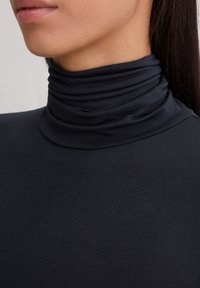 Nahaufnahme einer Person, die ein eng anliegendes schwarzes Rollkragenoberteil trägt. Die Hals-, Kieferlinie und Teile der Lippen sind sichtbar, ebenso wie das glatte, dunkle Haar.