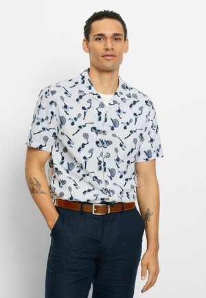 Mann trägt ein weißes Kurzarmhemd mit blauen Tennisspieler-Mustern, marineblaue Hose, braunen Gürtel und sichtbare Arm-Tattoos, steht mit einer Hand in der Tasche.