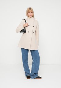 Cappotto doppiopetto in misto lana beige con colletto alto e grandi bottoni neri, abbinato a jeans in denim blu a gamba larga e scarpe nere.