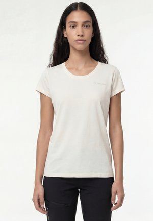 Giovane donna con lunghi capelli scuri che indossa una semplice t-shirt bianca a maniche corte e pantaloni neri, rivolta verso l'obiettivo su uno sfondo bianco.