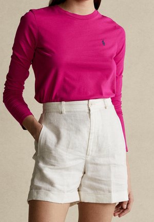 Vrouw die een felroze top met lange mouwen draagt, ingestopt in een hoogtailleshort van off-white linnen, met één hand in haar zak.