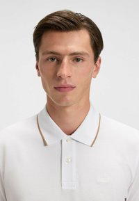 Vit pikétröja med en brun krage, med en knappslå och en broderad "BOSS"-logotyp på bröstet. Slät textur.