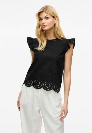 Top de algodón negro con mangas cortas con volantes y un dobladillo ondulado cortado a láser con patrones florales. Llevado con pantalones blancos de corte holgado.