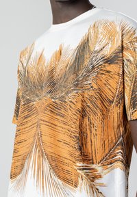 Short-sleeve t-shirt featuring an orange and brown palm leaf print on a white fabric background, with a round neckline and relaxed fit.