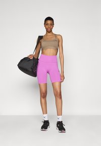 Un soutien-gorge de sport marron clair et un short de vélo rose vif, associés à des baskets noires et un sac de sport noir, sur fond blanc.