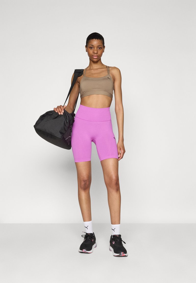 Un reggiseno sportivo marrone chiaro e pantaloni corti da bicicletta rosa brillante, abbinati a sneakers nere e a una borsa da palestra nera, il tutto su uno sfondo bianco.