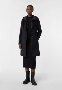 Cappotto nero con cappuccio, due bottoni e tasche laterali, indossato sopra un vestito nero e abbinato a stivali neri al ginocchio. Tessuto liscio.