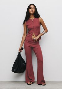Rúrový pletený dvojdielny outfit v ružovej farbe, s topom bez rukávov so zapínaním na gombíky a rozšírenými nohavicami. Drží čiernu tašku s očkovými detailmi.