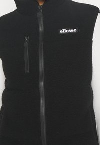 Maglione in pile nero con zip frontale completa, tasca sul petto con zip verticale e applicazione del logo "ellesse" bianco sul lato sinistro del petto.
