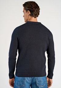 Marineblauer gerippter Strickpullover mit langen Ärmeln und rundem Ausschnitt, aus strukturiertem Stoff mit seitlichen Schlitzen, kombiniert mit blauen Jeans.