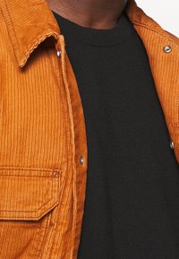 Rustfarbendes Kordhemd mit sichtbarer gerippter Textur und silbernen Druckknöpfen, kombiniert mit einem schwarzen Rundhalsausschnitt-Pullover darunter.