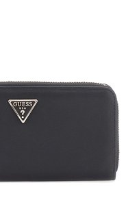 Portefeuille noir zippé en matière synthétique lisse, avec un logo triangulaire métallique proéminent et des détails de couture contrastés.