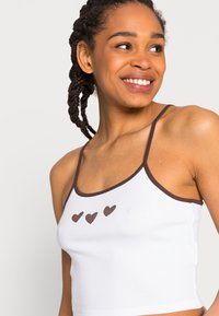 Top cropped branco rendado com detalhes castanhos e alças finas, apresentando três pequenos corações castanhos na frente. Textura suave.