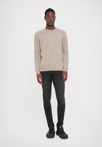 Uomo in piedi, che indossa un maglione lavorato a maglia beige, jeans neri slim e sneakers nere, su sfondo bianco uniforme.