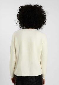 Maglione di lana lavorato a maglia di colore crema con maniche lunghe, scollo tondo e orlo a coste. Indossato sopra una gonna nera con texture, realizzata in un tessuto liscio e caldo.