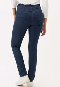 Jeans in denim blu scuro con una vestibilità slim, caratterizzati da una vita alta e due tasche posteriori, mostrati da un angolo posteriore. Sneakers bianche visibili.