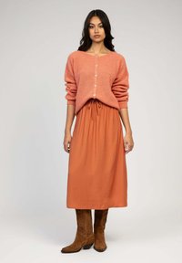 Cardigan côtelé rose avec boutons, associé à une jupe midi taille à nouer couleur rouille et des bottes de cowboy brodées marron. Texture lisse dans l'ensemble.