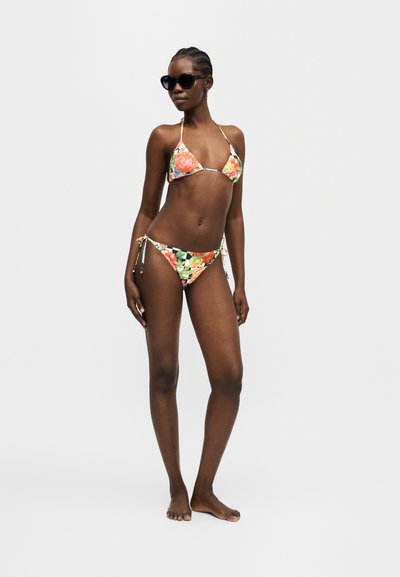 Γυναίκα με μπικίνι με λουλουδάτο σχέδιο και δεσίματα στο πλάι και μαύρα γυαλιά ηλίου, στέκεται ξυπόλυτη σε λευκό φωτεινό φόντο.