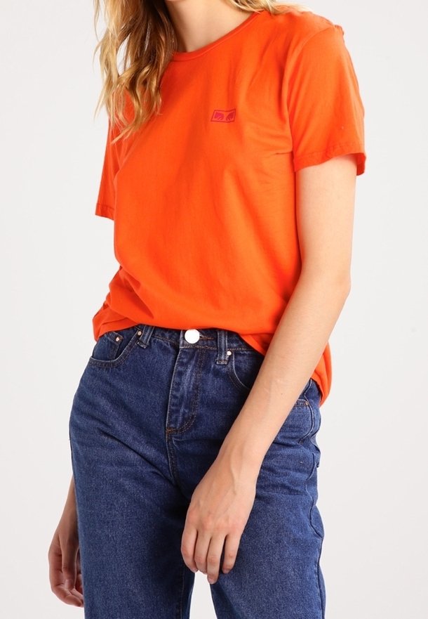 Camiseta de algodón naranja con cuello redondo y mangas cortas, que presenta un pequeño logo morado en el lado izquierdo del pecho. Combinada con pantalones vaqueros de cintura alta en azul.