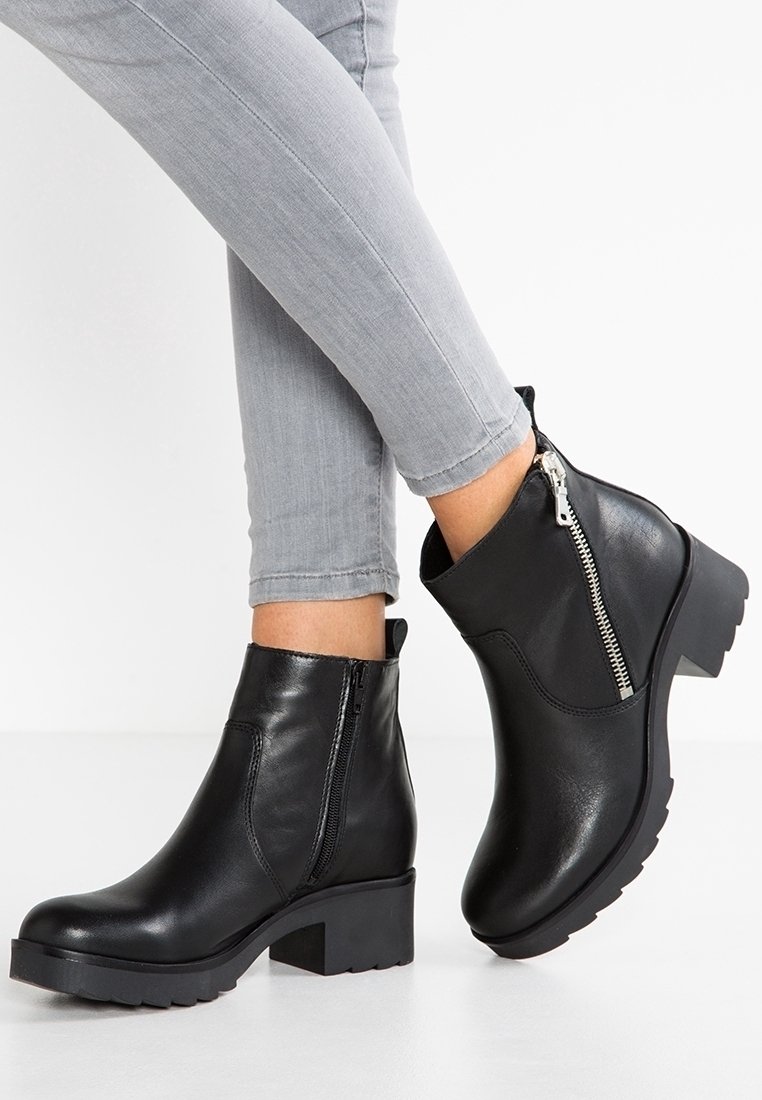Bottes noires en cuir avec un talon épais, fermeture éclair latérale et semelle texturée, portées avec un jean gris slim.