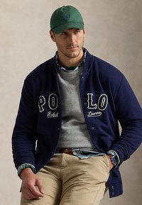 Άνδρας με πράσινο καπέλο, μπλε σκούρο μπουφάν "Polo Ralph Lauren" πάνω από γκρι φούτερ και πουκάμισο με καρό, μπεζ παντελόνι, καθιστός με το χέρι στην τσέπη.