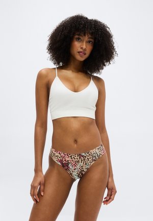 Γυναίκα με σγουρά μαλλιά που φοράει λευκό μπραλέτα και πολύχρωμο εσώρουχο με λεοπάρ σχέδιο, σε απλό ανοιχτό φόντο.