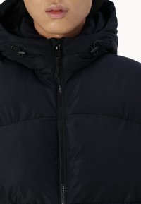 Czarna pikowana kurtka z wysokim kołnierzem i regulowanym kapturem. Posiada centralny zamek błyskawiczny oraz teksturowany materiał. Charakterystyczne przeszycia widoczne na klatce piersiowej.