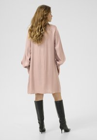 Vestido de manga larga de color rosa claro con una textura suave, que presenta mangas abullonadas y un corte relajado, combinado con botas altas negras de tacón de aguja.