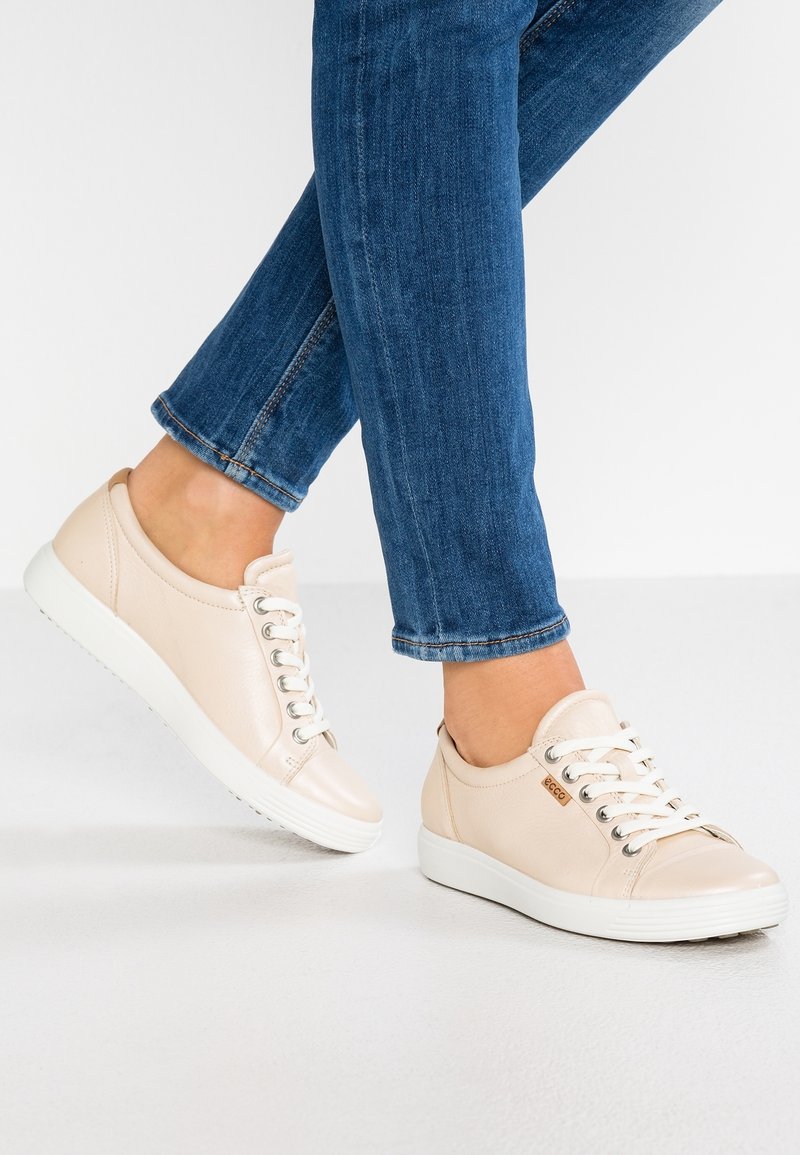 Beige leather sneakers with white rubber soles, round toe, and flexible design. Features silver eyelets and lace closure. Earthy color scheme.