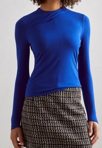 Blå långärmad topp med draperad halsringning, som har en mjuk textur. Kombinerad med en svart- och vitmönstrad kjol.