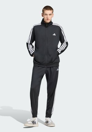 Junger Mann steht mit den Händen in den Taschen, trägt einen schwarzen Adidas-Trainingsanzug mit weißen Streifen und weißen Sneakers vor schlichtem Hintergrund.