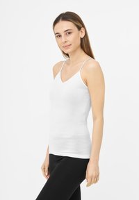 Caraco blanc à encolure en V en tissu lisse avec fines bretelles, coupe ajustée et ourlet sans couture. Associé à des leggings noirs.