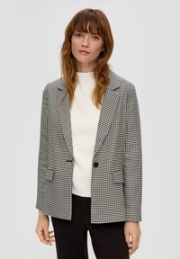 Schwarzweißes Hahnentrittmuster-Blazer mit einem Reverskragen, einem Knopfverschluss und zwei Fronttaschen. Über einem weißen Oberteil getragen.