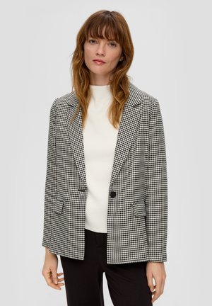 Blazer a scacchi bianco e nero con colletto a punta, chiusura a singolo bottone e due tasche frontali. Indossato sopra una maglietta bianca.