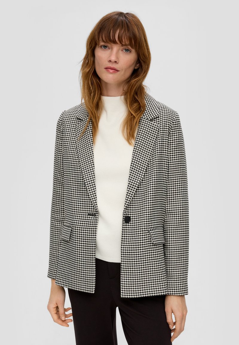 Schwarzweißes Hahnentrittmuster-Blazer mit einem Reverskragen, einem Knopfverschluss und zwei Fronttaschen. Über einem weißen Oberteil getragen.