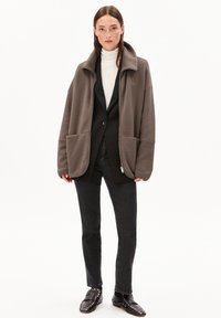 Femme portant une veste en polaire taupe avec de grandes poches, un blazer noir, un col roulé blanc, un jean noir et des chaussures à clous noires, se tenant contre un fond blanc.