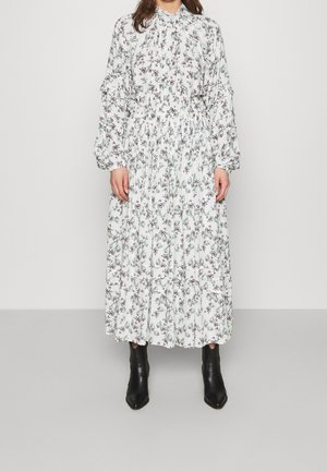 Woman wearing a long, white floral dress with long puffed sleeves and black ankle boots, standing against a plain background.