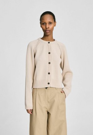 Femme portant un cardigan beige boutonné et un pantalon large kaki, debout avec une main dans la poche devant un fond uni.